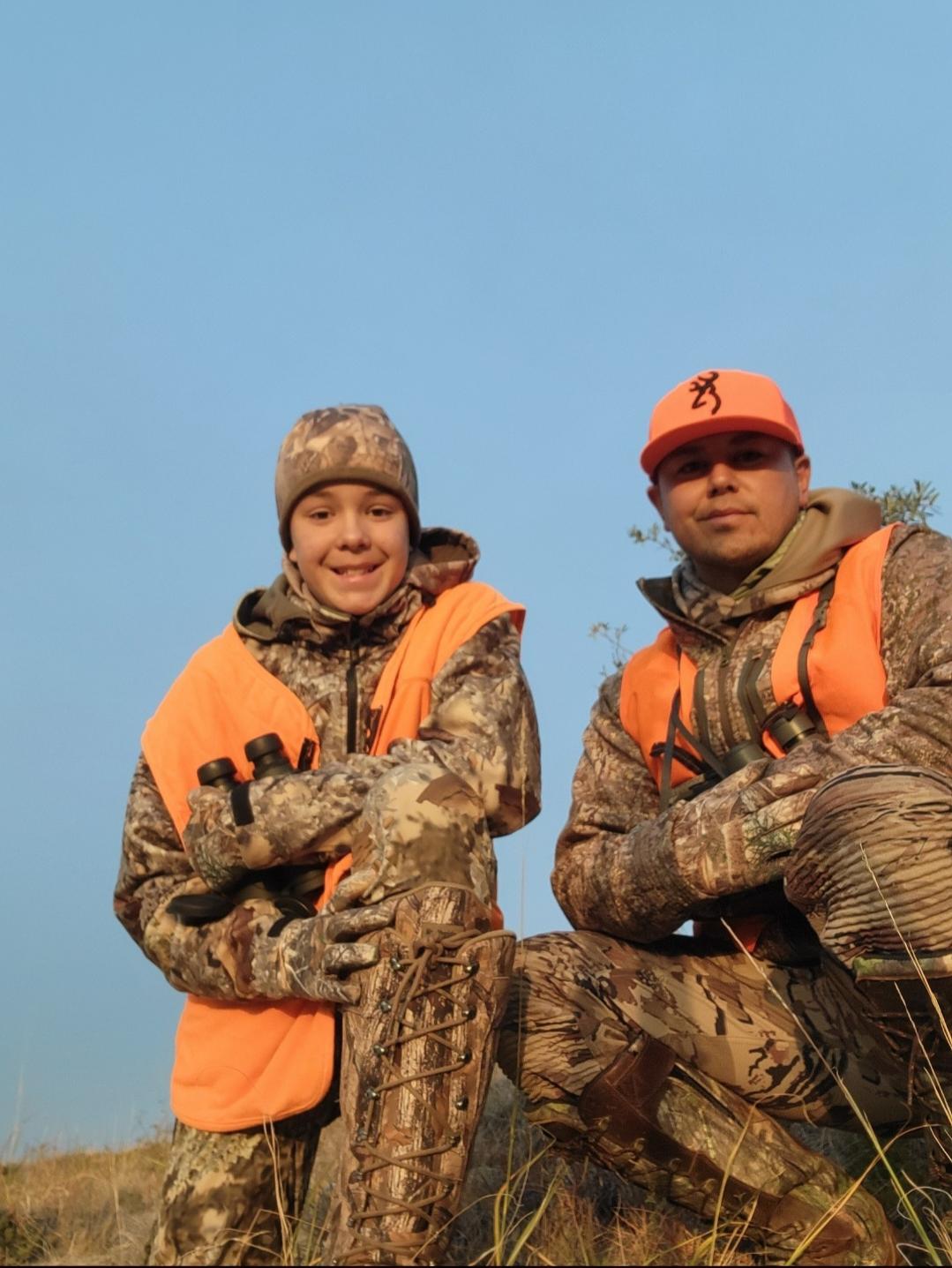 A man and a child, both wearing orange hunting vests and camouflage gear, are kneeling on grassy terrain.